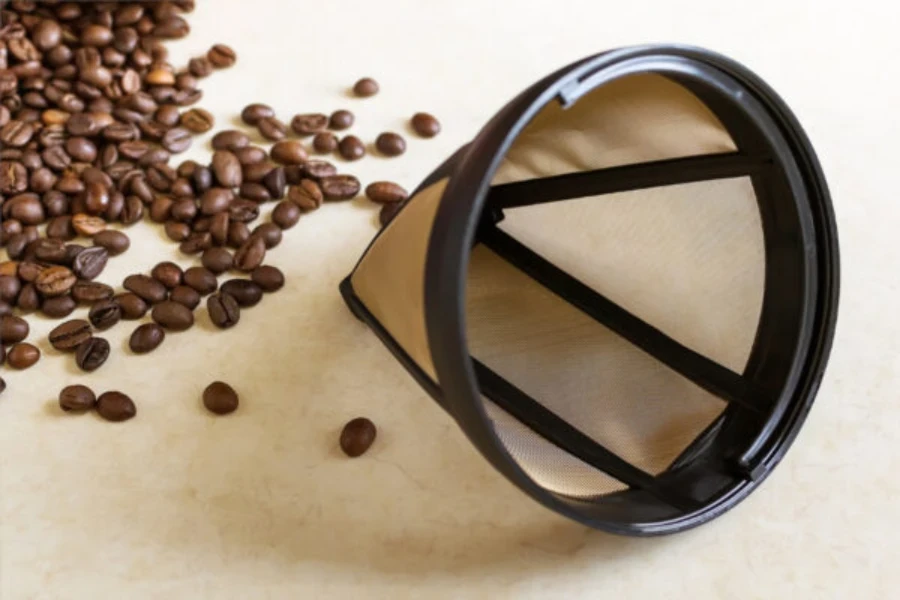 A reusable metal mesh coffee filter on a surface next to a pile of coffee beans