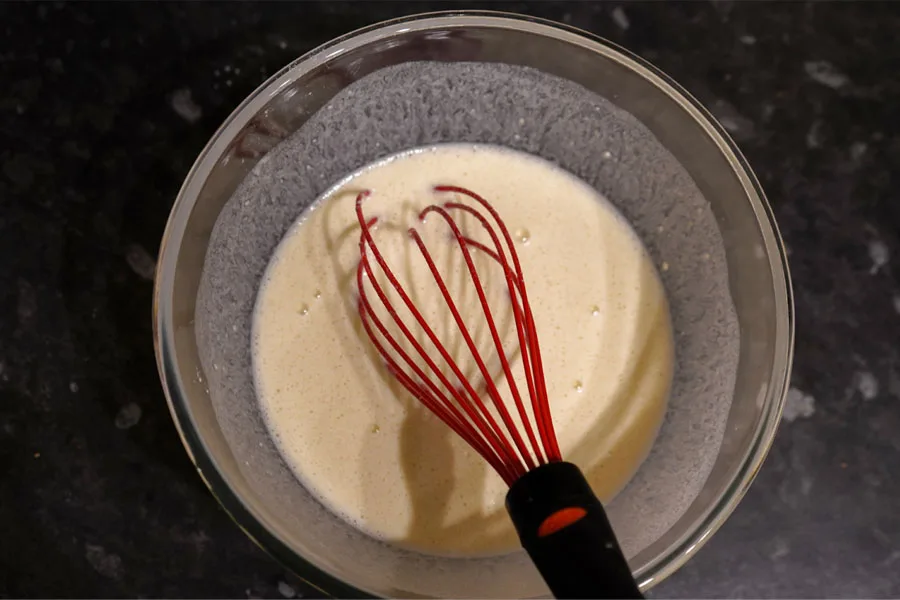 A glass mixing bowl with batter and a whisker