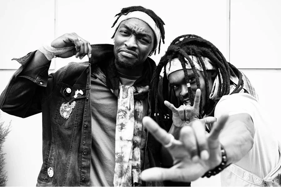 Black and white young black man frowning and pulling up collar of stylish denim coat while cheerful friend demonstrating sign of horns and looking at camera