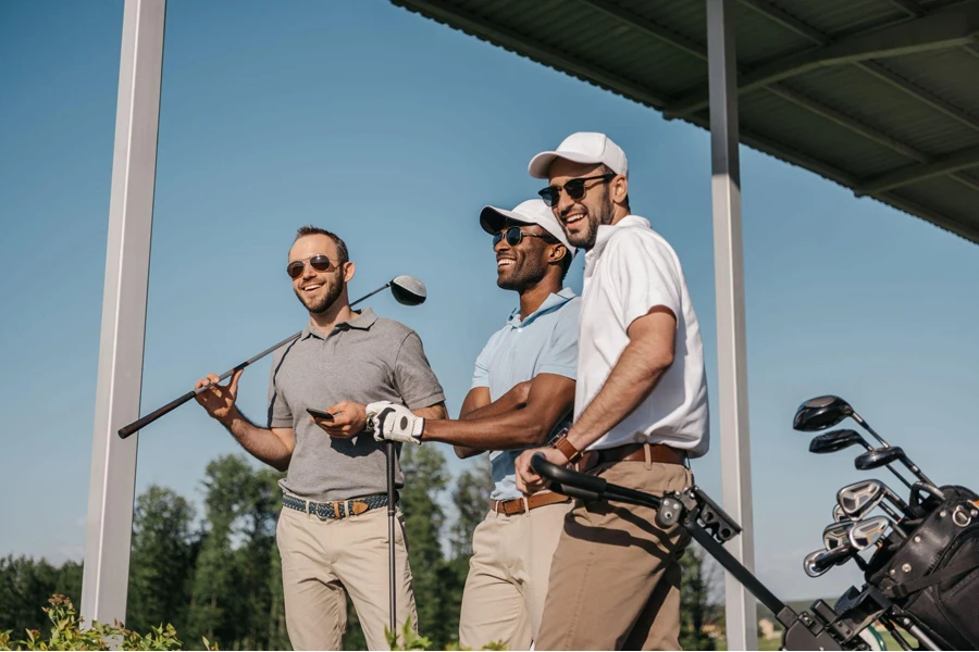 Tiga pria tersenyum berkacamata hitam memegang tongkat golf di luar ruangan