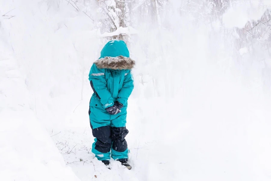 フード付きのターコイズブルーのオーバーオールを着た少年が雪の森に立ち、雪が彼の上に降り注ぐ