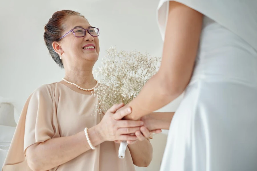 Счастливая пожилая женщина трогает руки дочери, стоящей в свадебном платье, готовой к церемонии