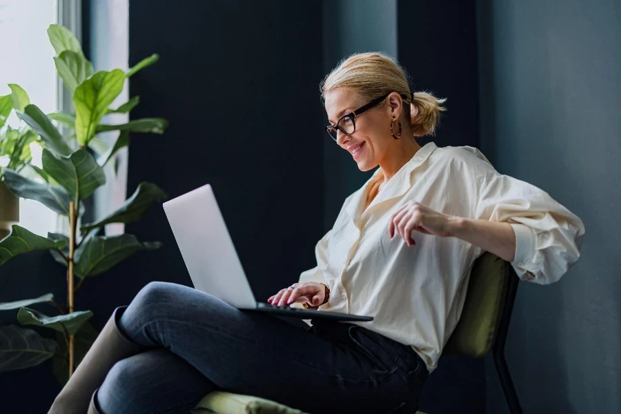 Alegre mujer de negocios sonriente sentada en una silla y escribiendo un informe de negocios en un teclado de computadora portátil