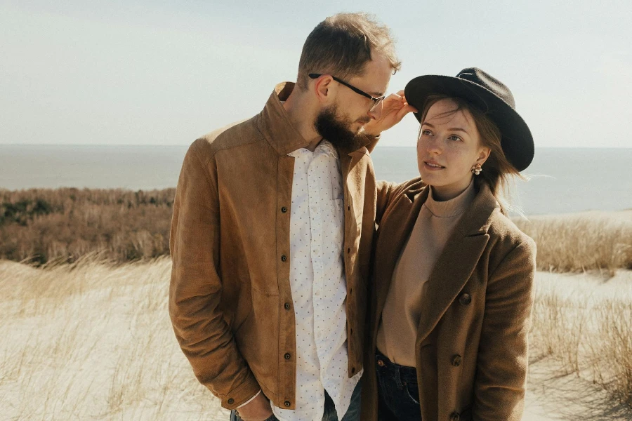 Homem com jaqueta de couro e mulher com casaco marrom e chapéu Fedora posando em uma duna