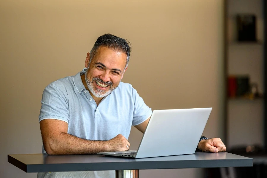 Porträt eines glücklichen, reifen Geschäftsmannes, der an einem Tisch steht und an seinem Laptop arbeitet und in die Kamera blickt