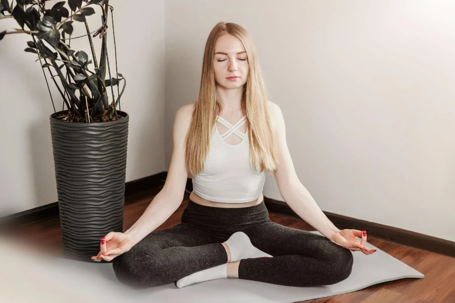 Blonde Frau sitzt mit geschlossenen Augen auf einer Yogamatte und meditiert zu Hause