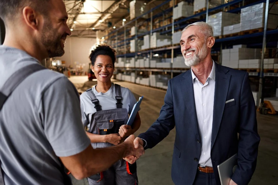 Happy manager and manual worker shaking hands next to black female colleague in distribution warehouse
