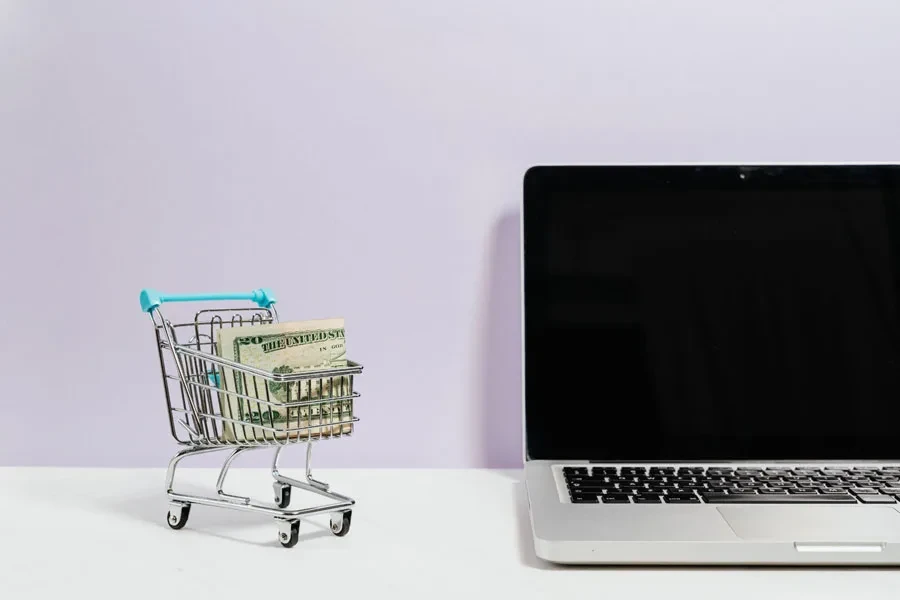 macbook pro sobre una mesa blanca junto a un carrito de compras en miniatura con dinero