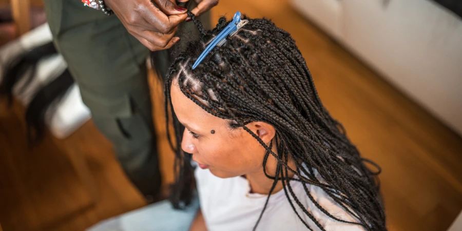 Профессиональная чернокожая женщина-парикмахер среднего возраста старательно работает над заплетением волос клиентки смешанной расы в домашних условиях.