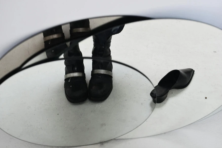 Beschneiden Sie die Füße einer anonymen Person in schwarzen Stiefeln mit dekorativen Metalldetails, die neben einem schwarzen, stylischen Schuh stehen und sich im ovalen Spiegel spiegeln