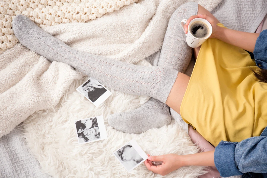 Modisch gekleidetes Mädchen sitzt auf dem Bett und trinkt Kaffee, während es sich Fotos ansieht