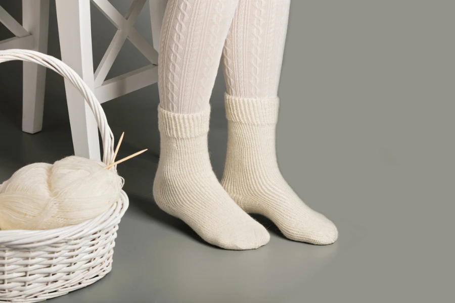 Piedi femminili in calze e calzini lavorati a maglia bianchi vicino al cestino con filato e maglia