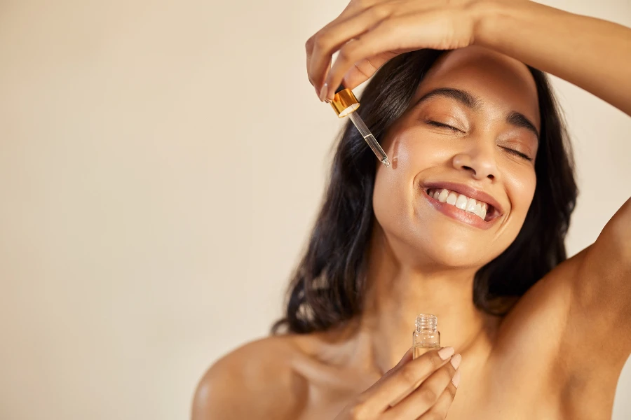 Happy young woman with bare shoulder applying serum on face with closed eyes