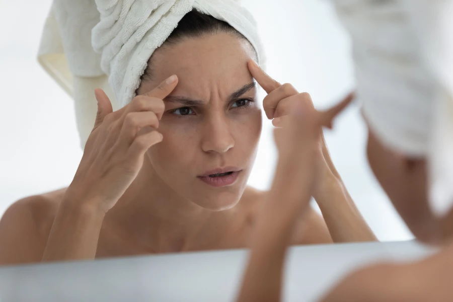 Обеспокоенная молодая женщина, завернутая в полотенце, смотрит в зеркало в ванной комнате