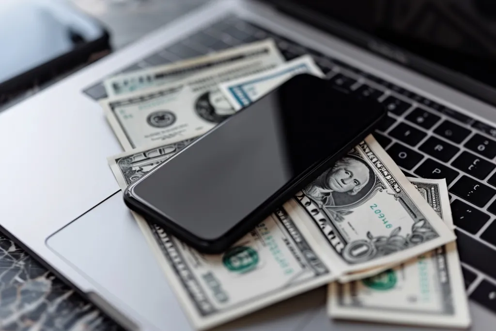 Sebuah telepon diletakkan di atas sejumlah uang tunai di samping komputer laptop yang terbuka