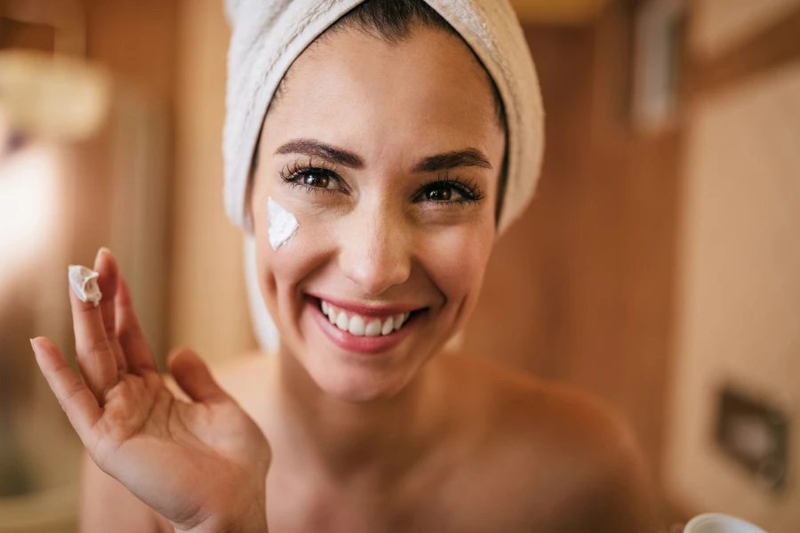 Linda mulher sorridente aplicando creme facial no banheiro