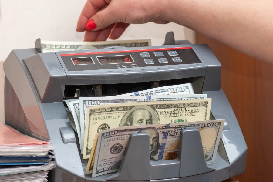 Woman cashier putting one hundred dollars American banknotes into currency counter at checkout counter