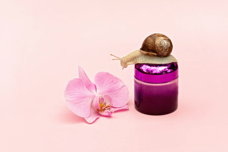 Cosmetici per il ringiovanimento della pelle su sfondo rosa con lumaca e fiori di orchidea
