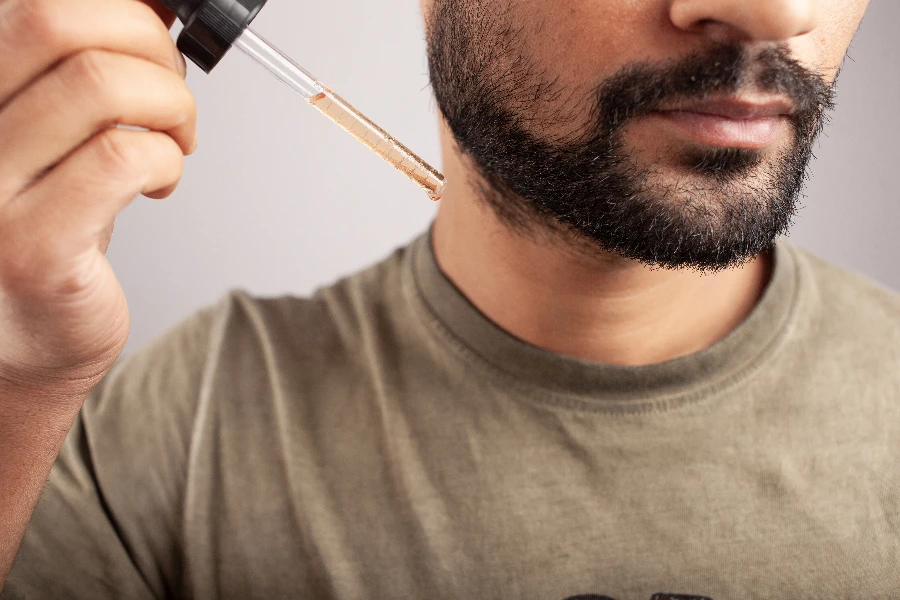 vista in primo piano di un uomo che tiene un contagocce pieno di minoxidil che va applicato per la crescita della barba isolato su sfondo bianco