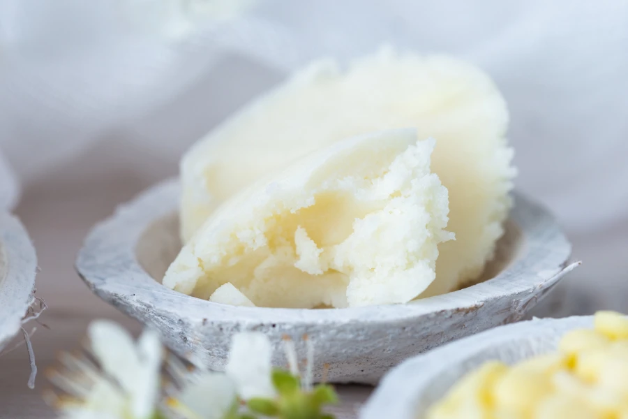 A selective focus shot of shea butter in a saucer and some white flowers on a white table top
