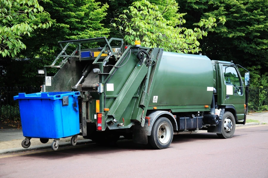 Truk sampah hijau dengan tempat sampah beroda biru yang ditinggikan di bagian belakang