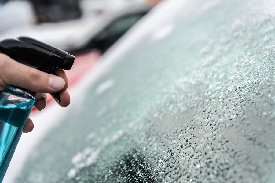 Mann taut mit einer Flasche Enteisungsmittel die vereiste Windschutzscheibe seines Autos auf