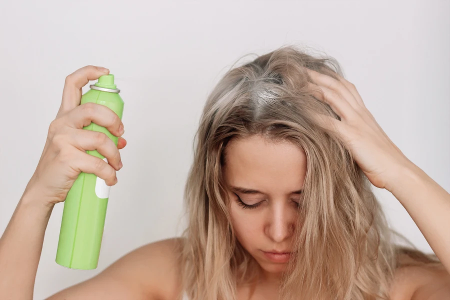 Junge blonde Frau mit schmutzigem fettigem Haar sprüht Trockenshampoo