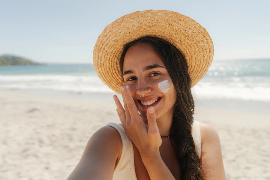 Foto de uma jovem aplicando protetor solar no rosto enquanto está na praia