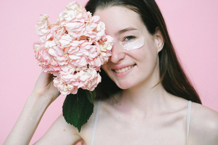 Femme souriante couvrant les yeux avec une fleur rose en se tenant debout sur fond rose