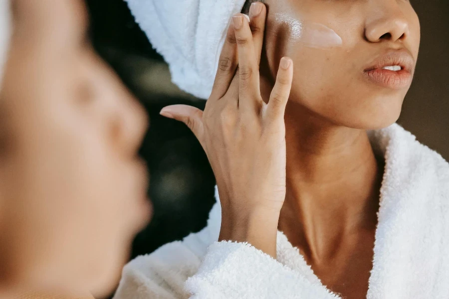 young ethnic female in terry robe applying CC cream on cheek while reflecting in mirror 