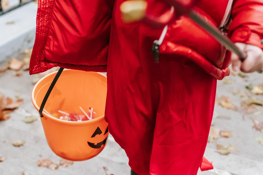 Pangkas anak anonim dengan kostum setan merah untuk Halloween dengan garpu rumput berjalan di jalan pada hari musim gugur