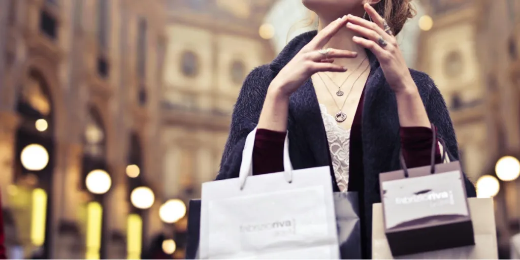 Женщина в очках, в сером пальто с глубоким вырезом и длинными рукавами и с фирменными бумажными пакетами.