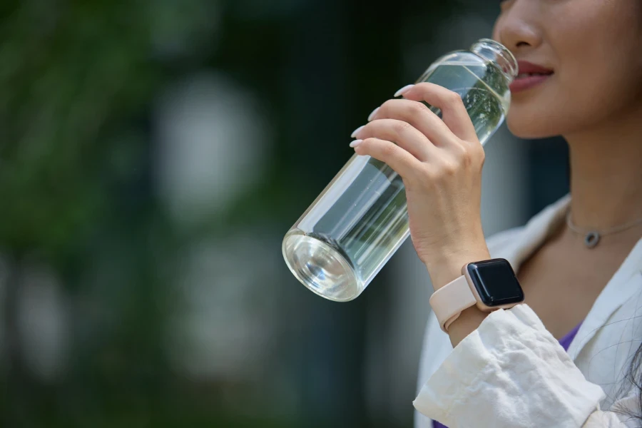 Giovane donna del sud-est asiatico fotografata nella vita quotidiana con uno smartphone moderno, cuffie wireless, una bottiglia d'acqua in vetro alla moda e una borsa di cotone sostenibile