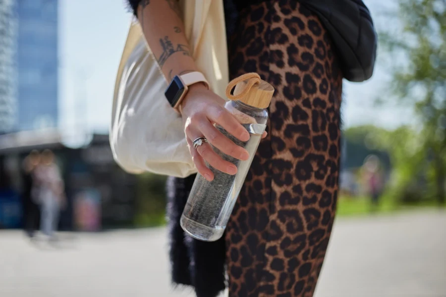 Nicht erkennbare weibliche Person hält eine wiederverwendbare Glasflasche mit frischem Wasser in der tätowierten Hand