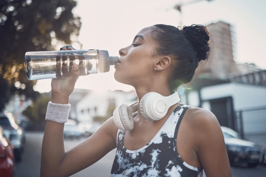 Joven mujer del Sudeste Asiático fotografiada en la vida diaria con un moderno teléfono inteligente, auriculares inalámbricos, una moderna botella de agua de vidrio y una bolsa de algodón sostenible