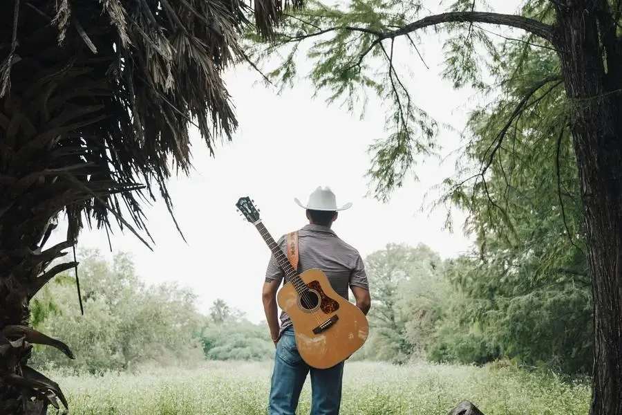 Un hombre con sombrero de vaquero sosteniendo una guitarra en el bosque por Eli Villarreal