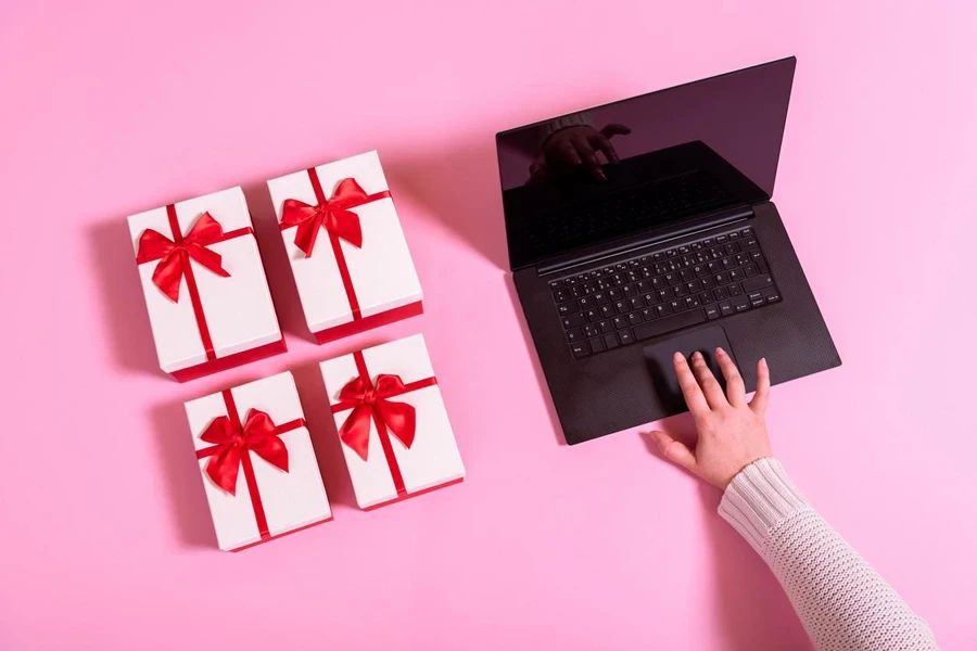 Sopra la vista con la mano di una donna che acquista regali online da un laptop. Commercio elettronico con un taccuino e scatole regalo su un tavolo rosa.