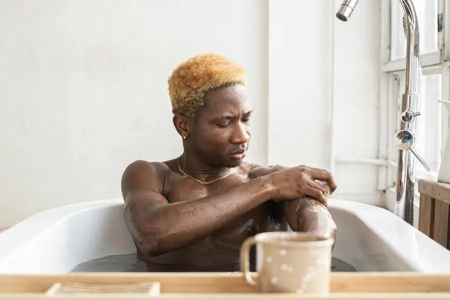 Homme afro-américain assis dans une baignoire avec une tasse de café sur un plateau et se frottant les bras avec une éponge et du savon par Monstera Production