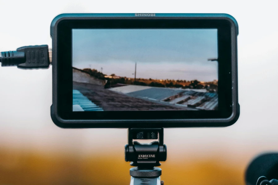 Contemporary rectangular shaped video camera monitor with charger and display showing pictures of roofs of houses in city under blue sky on tripod