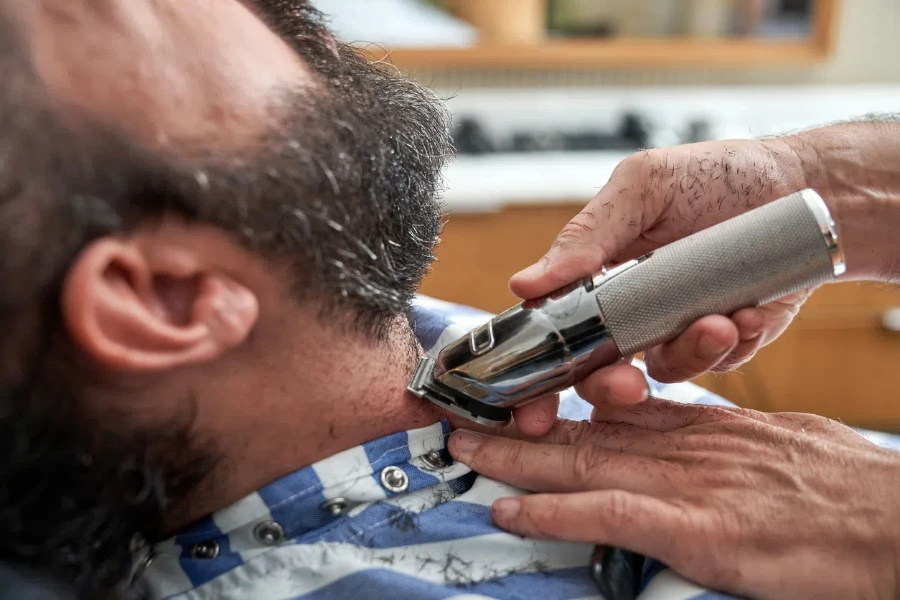 Anonymer männlicher Friseur schneidet einem bärtigen männlichen Kunden im Friseursalon die Haare mit einem Trimmer.