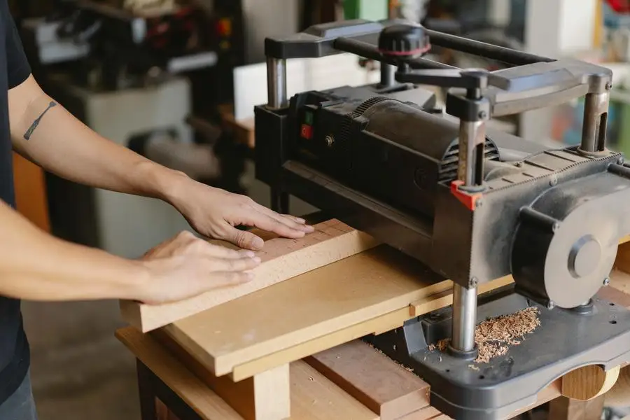 Crop anonymous tattooed woodworker leveling wooden plank on professional electric equipment in workshop in daylight by Ono Kosuki