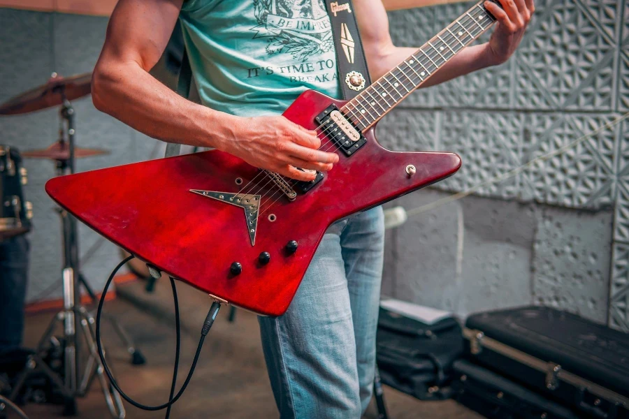 Pemuzik lelaki berbaju tee dan berseluar jeans berdiri dan bermain gitar elektrik merah dengan gendang di belakang