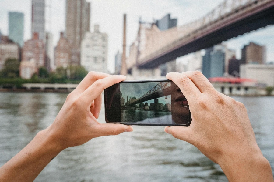 Ritaglia un viaggiatore irriconoscibile che scatta una foto del moderno ponte sospeso in città usando il cellulare durante il giorno