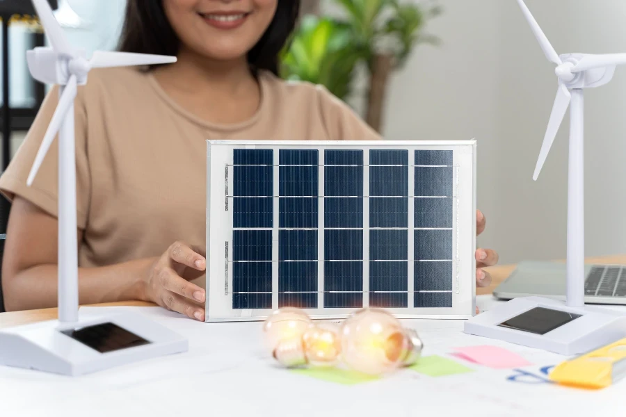 Vue recadrée d'une femme tenant une ampoule sur une table blanche avec un modèle d'éolienne, de cellule solaire et de blockhaus sur une table de travail