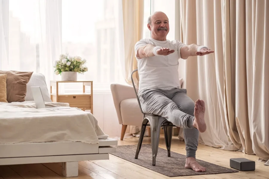 Пожилой мужчина практикует асаны йоги или спортивные упражнения для ног и рук на стуле