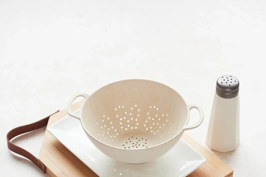 Empty colander on white plate and wooden cutting board with leather handle, next to the salt shaker on the table with copy space.