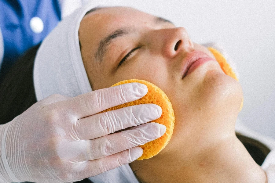 Female beautician in medical robe and gloves using yellow sponges