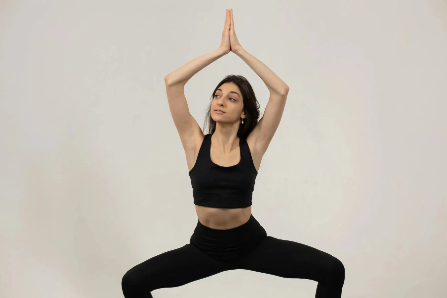 Fit female athlete in sportswear doing plie squat with namaste gesture above head against white background