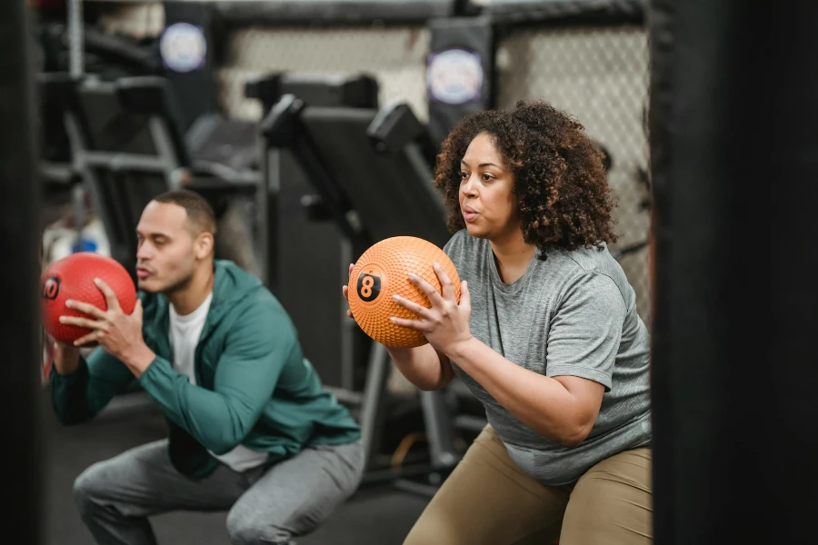 Mulher negra jovem plus size focada em roupas esportivas fazendo agachamentos com bola medicinal enquanto treina na academia com treinador muscular étnico masculino