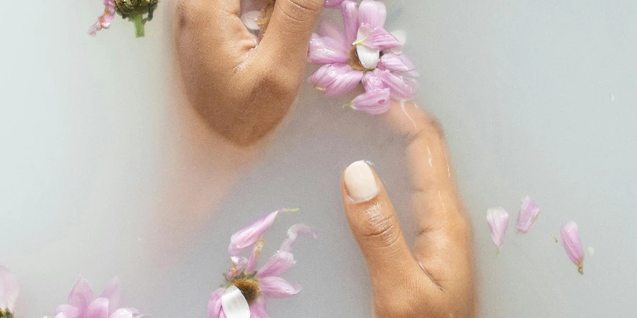 From above of anonymous female with manicure touching delicate petals of pink flowers in soap water